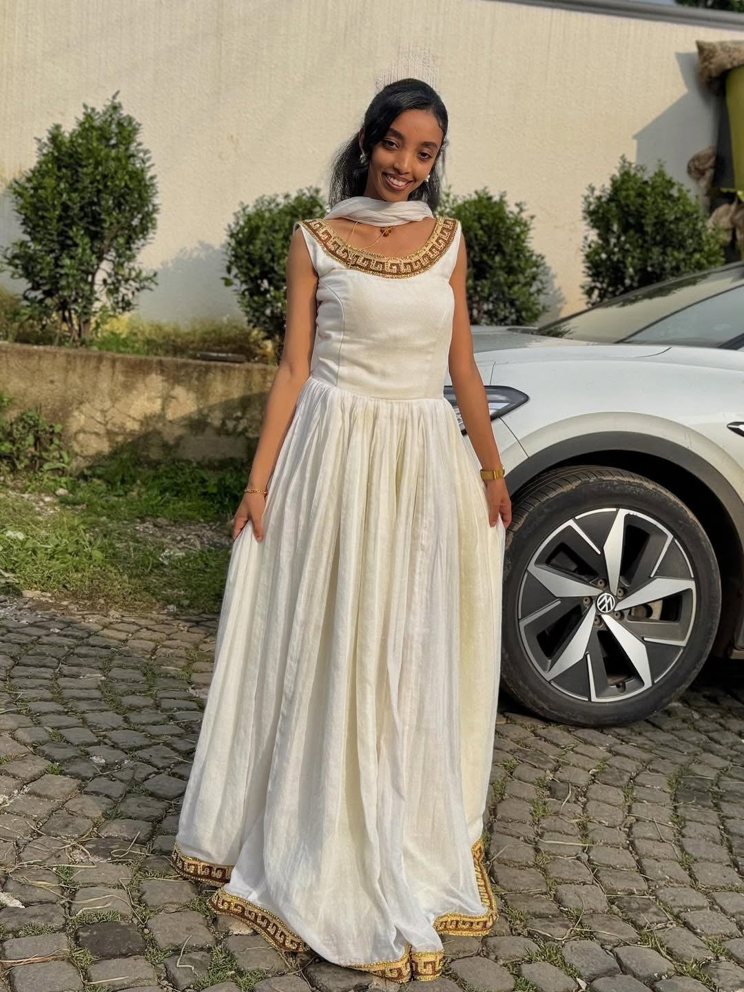 Woman in a Gold Ethiopian Dress with gold accents standing outdoors near a car.