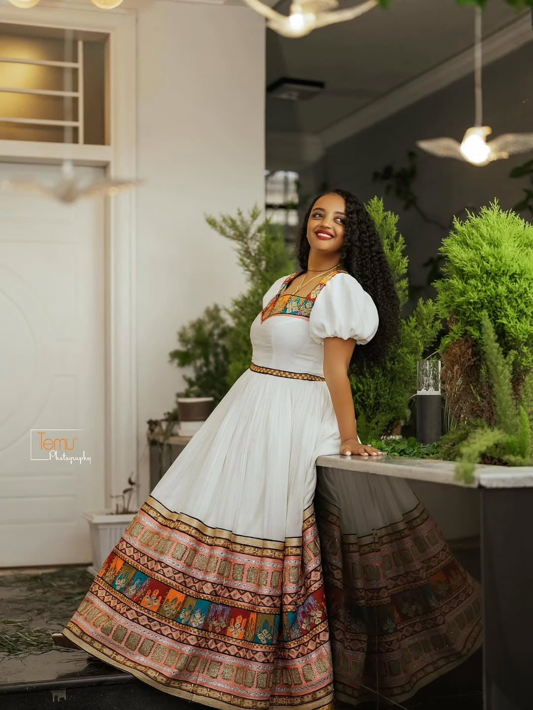 Woman in a Brown Habesha Zuria with colorful patterns standing in an indoor setting with plants.