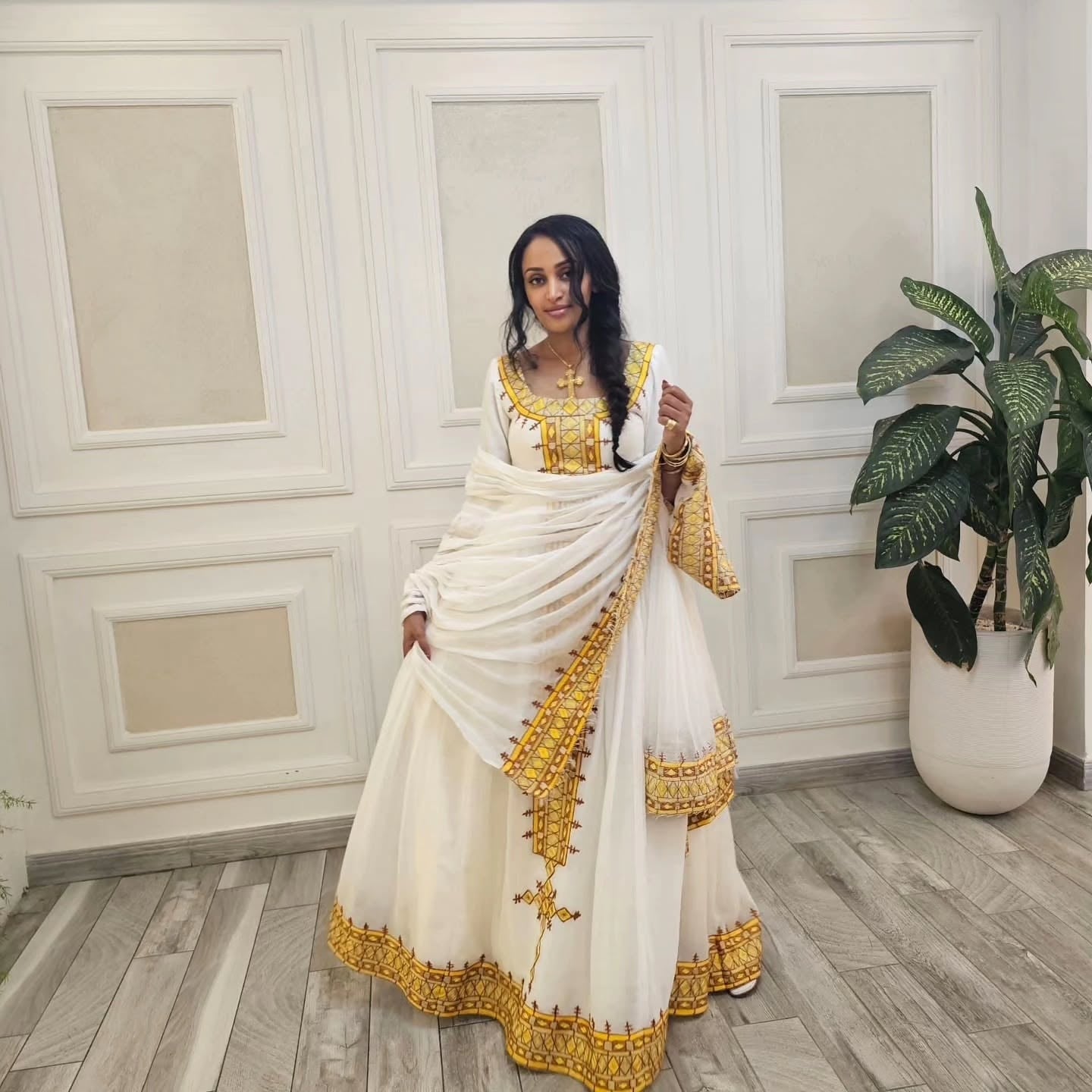 Woman in a white and gold traditional habesha dress standing in a room with decorative walls and a plant.