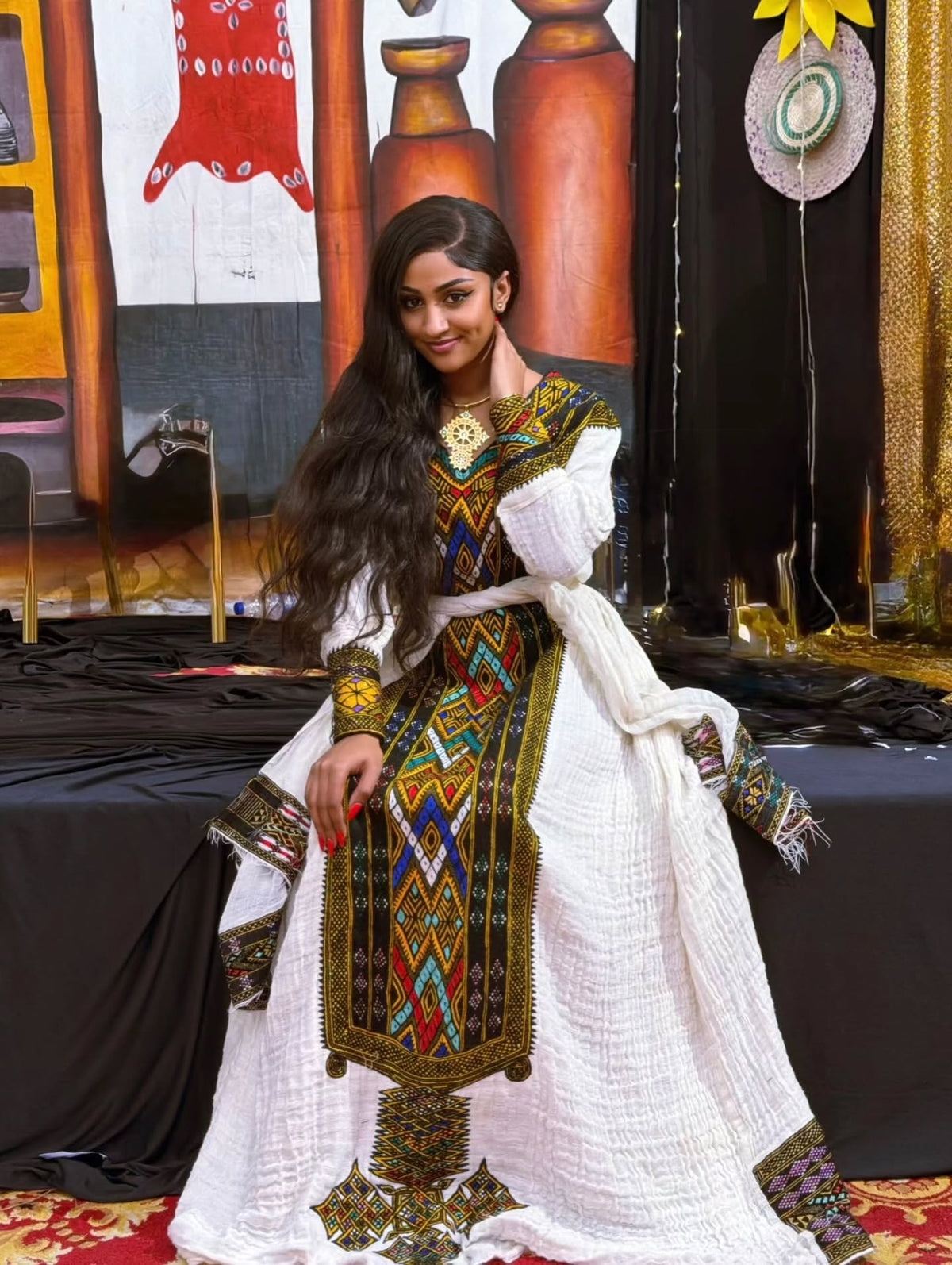 A woman models a traditional white embroidered Gonder fetel  with colorful geometric patterns and a white textured shawl, set against a background of draped fabrics and decorative elements.