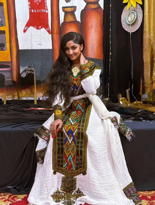 A woman models a traditional white embroidered Gonder fetel  with colorful geometric patterns and a white textured shawl, set against a background of draped fabrics and decorative elements.