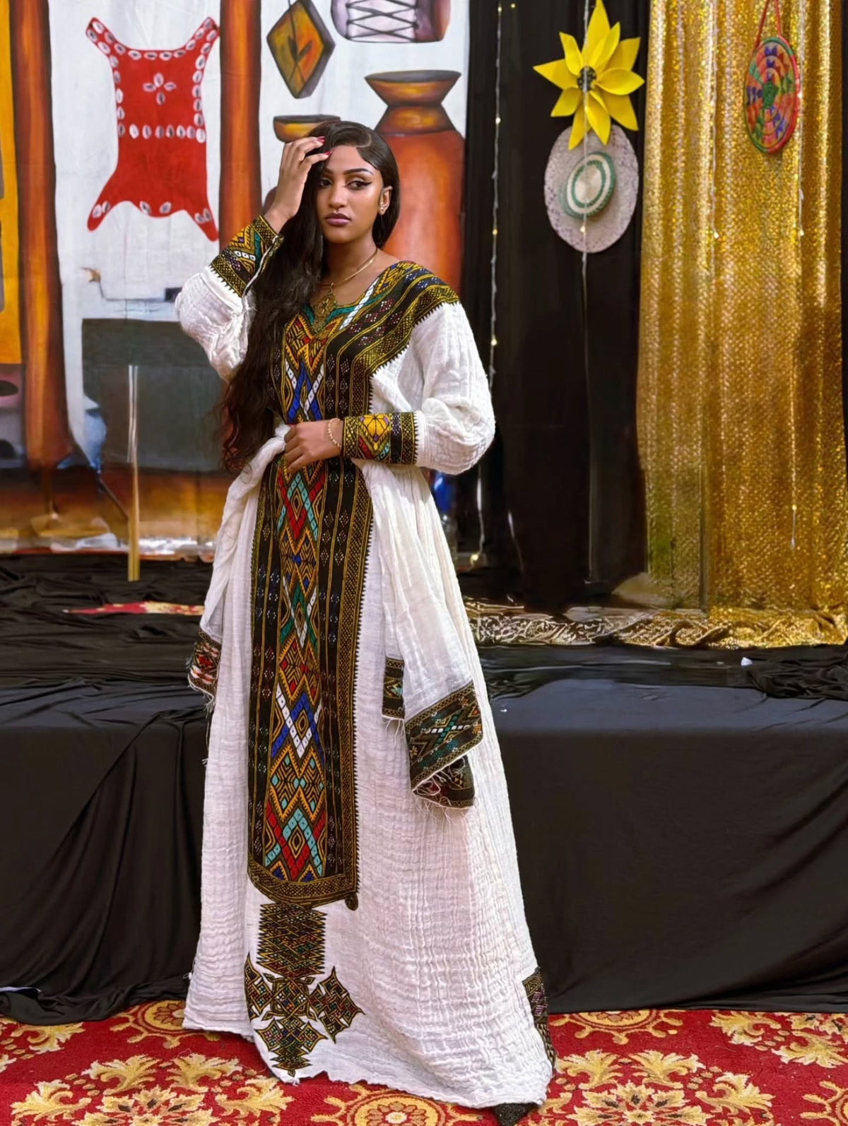 A woman modeling a traditional white long Gonder fetel with intricate multicolor geometric embroidery and beadwork, standing on a red patterned carpet in front of a decorated stage backdrop.