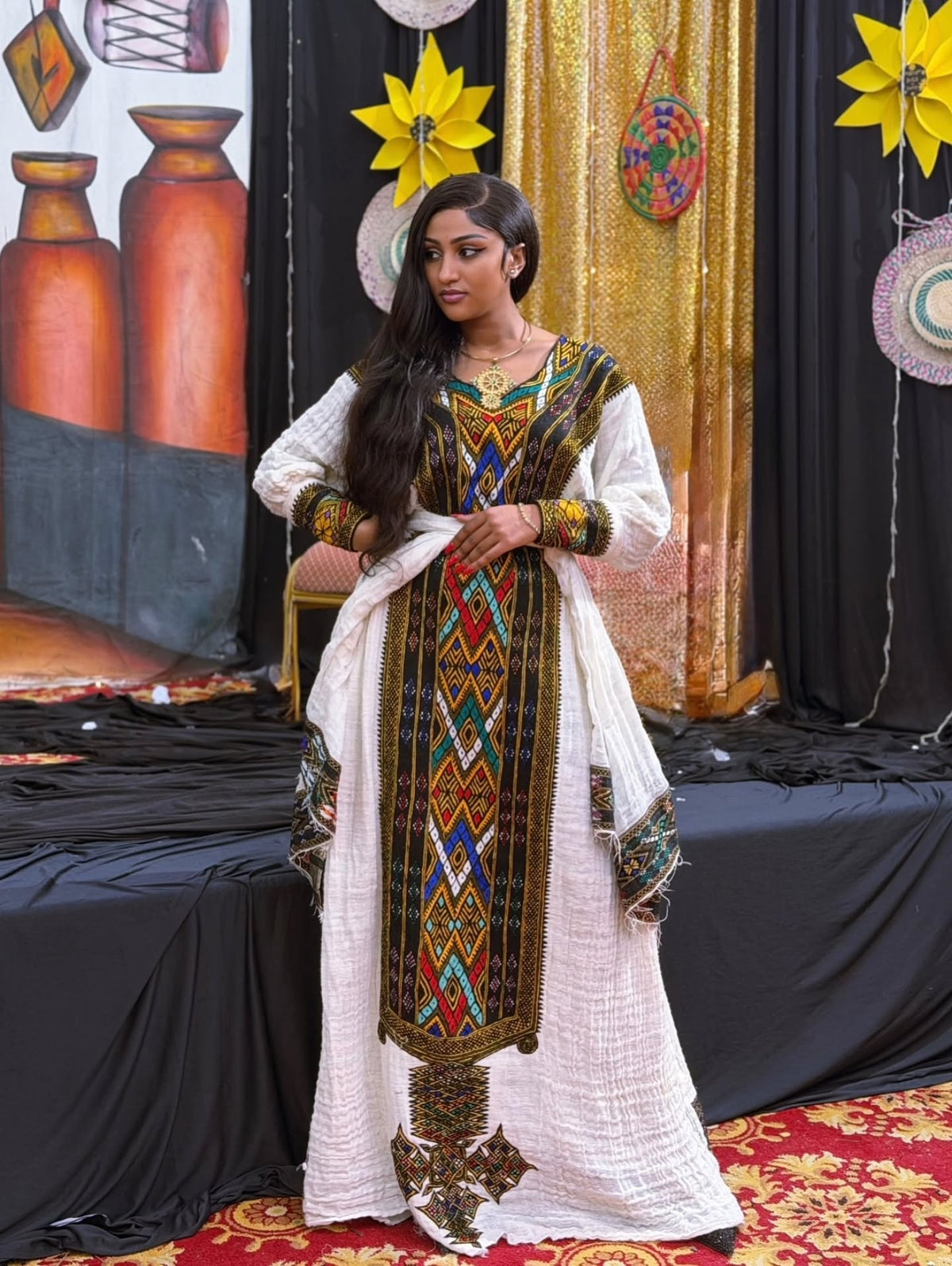 A woman models a traditional white Gonder fetel  kurta with a matching dupatta and a highly detailed multicolor geometric embroidered panel down the front. The setting is a decorated stage with black drapes, gold fabric, and decorative paper stars.