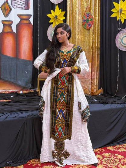 A woman models a traditional white Gonder fetel  kurta with a matching dupatta and a highly detailed multicolor geometric embroidered panel down the front. The setting is a decorated stage with black drapes, gold fabric, and decorative paper stars.