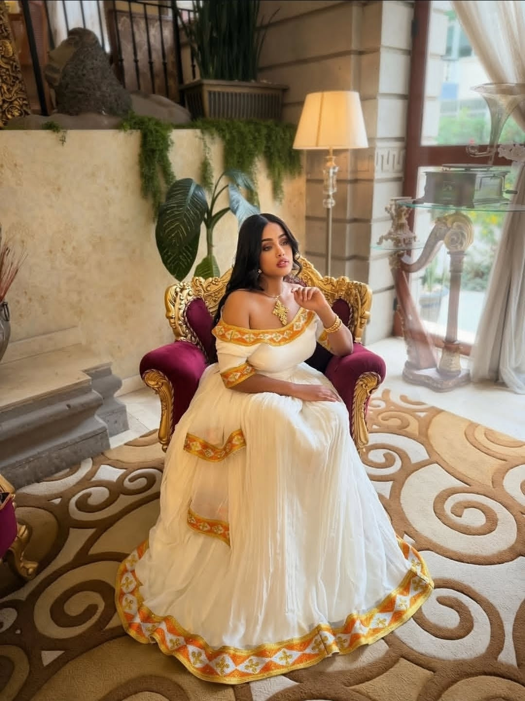Woman in a white and gold habesha dress sitting on a decorative chair in an elegant interior setting.