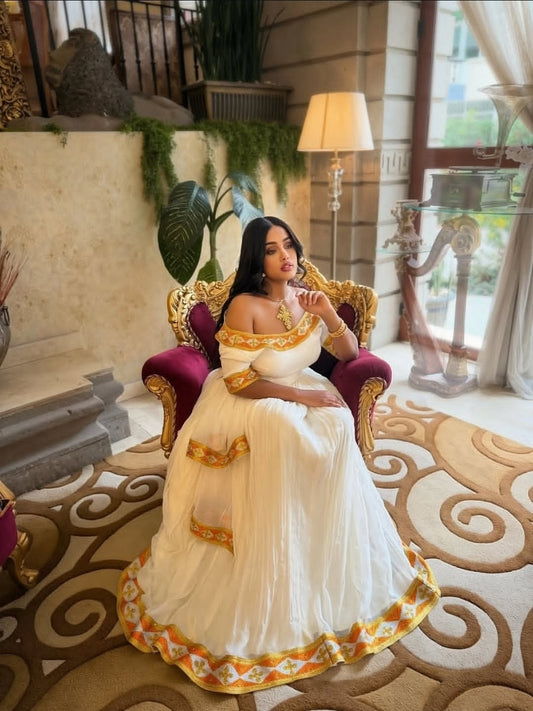 Woman in a white and gold habesha dress sitting on a decorative chair in an elegant interior setting.