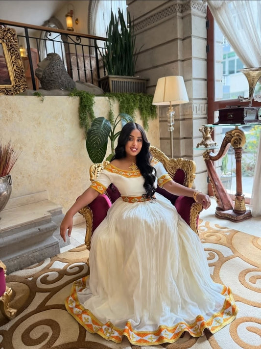 Woman in a white and gold habesha dress sitting on a decorative chair in an elegant interior setting.