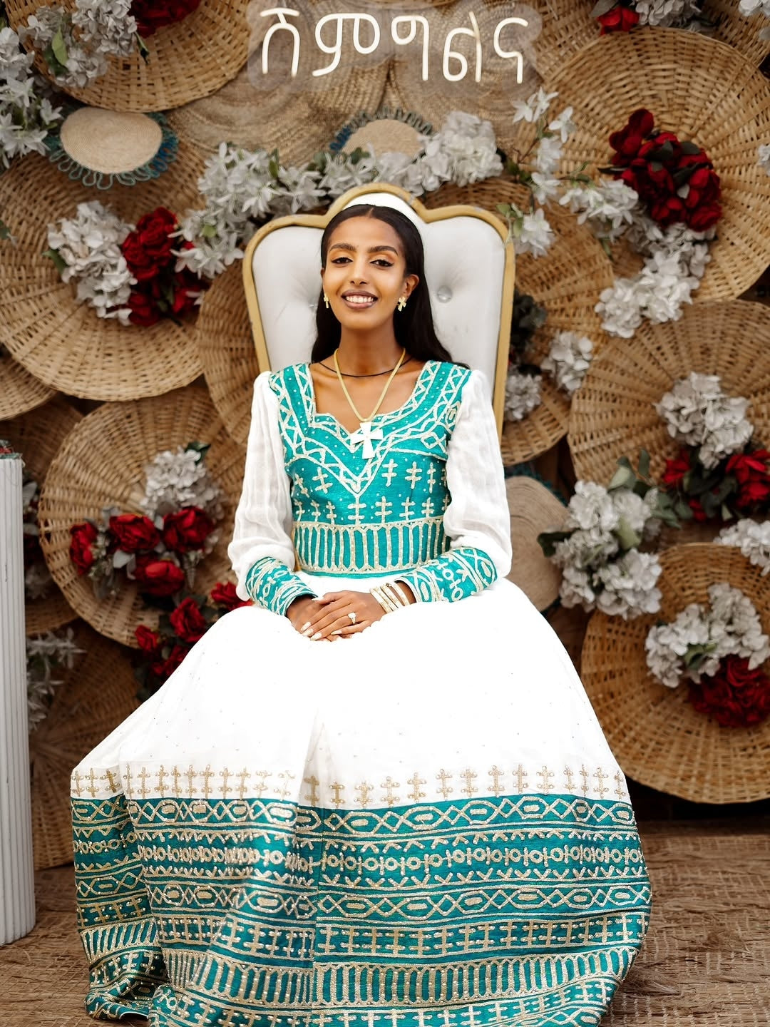 Woman in Ethiopian wedding attire sitting on a decorative chair with floral and hat decorations.
