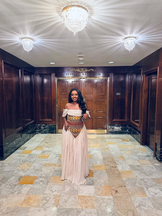 Woman in a long, patterned habesha dress standing in a formal setting with wooden doors and chandeliers.