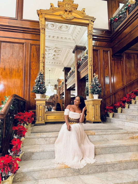 Woman in a white habesha dress standing on a staircase decorated with poinsettias and a large mirror.