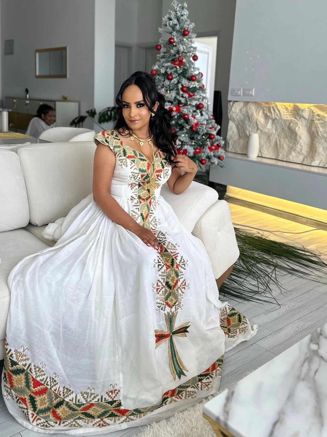 Woman in a white Ethiopian libs with colorful patterns sitting on a couch in a modern living room with a Christmas tree.