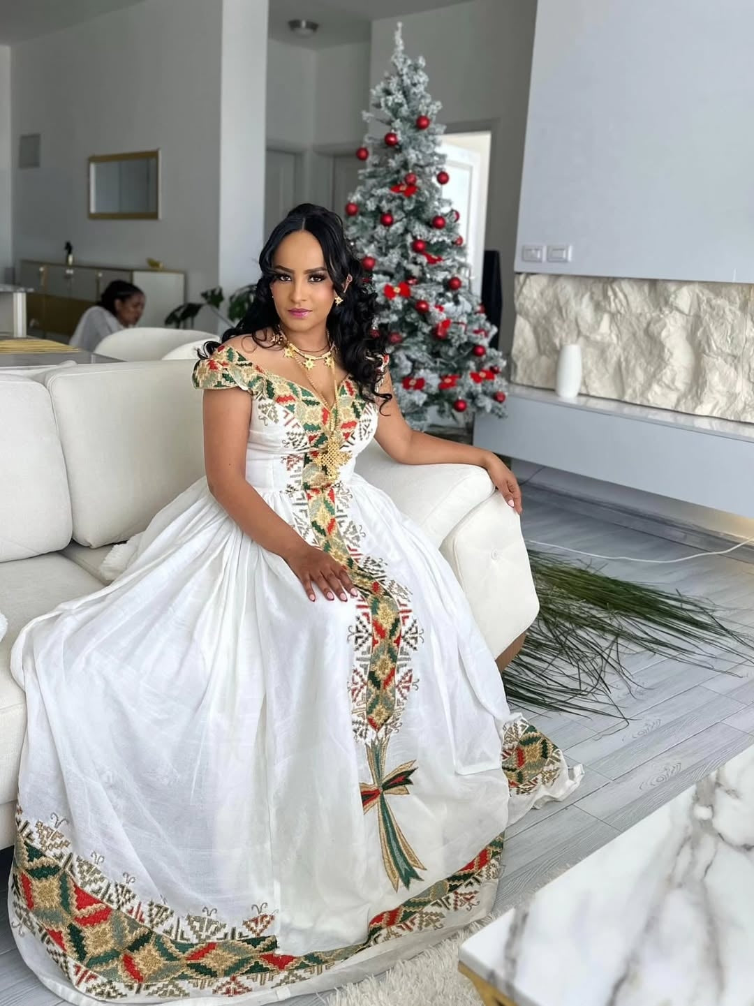 Woman in a white Ethiopian libs with floral patterns sitting in a modern living room with a Christmas tree.