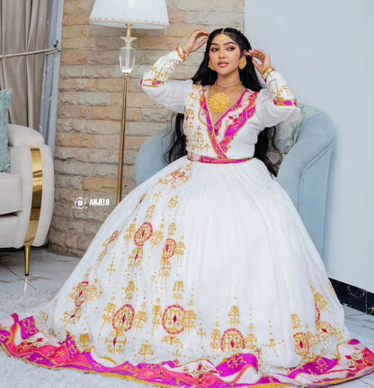 Woman in a white  embroidered Ethiopian Zuria  with pink and gold accents sitting on a couch.
