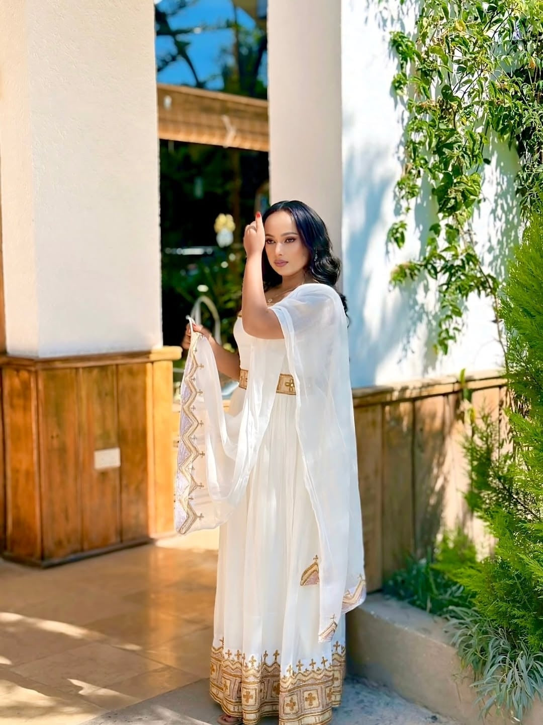 Woman in a white and gold Habesha Kemis standing outdoors with greenery and a building in the background.