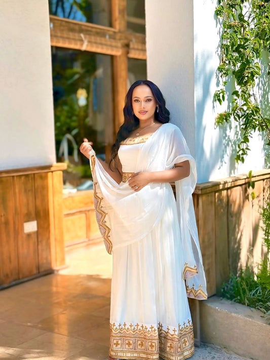 Woman in a white Habesha Kemis with gold patterns standing outdoors.