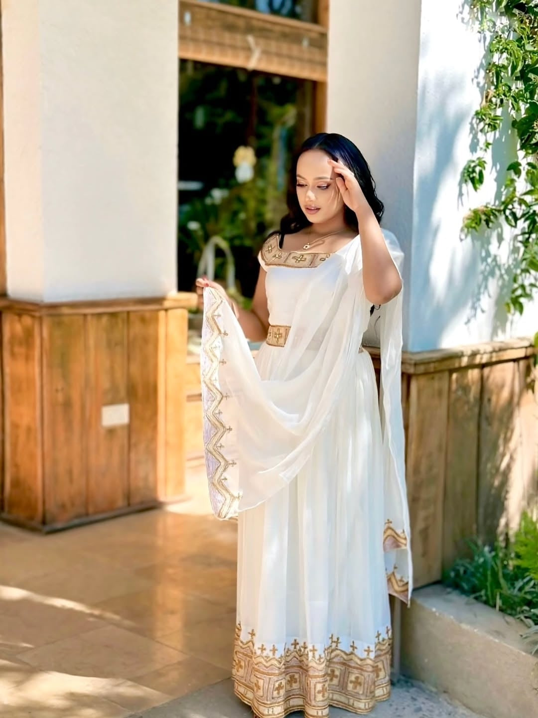 Woman in a white Habesha Kemis with gold patterns standing outdoors.
