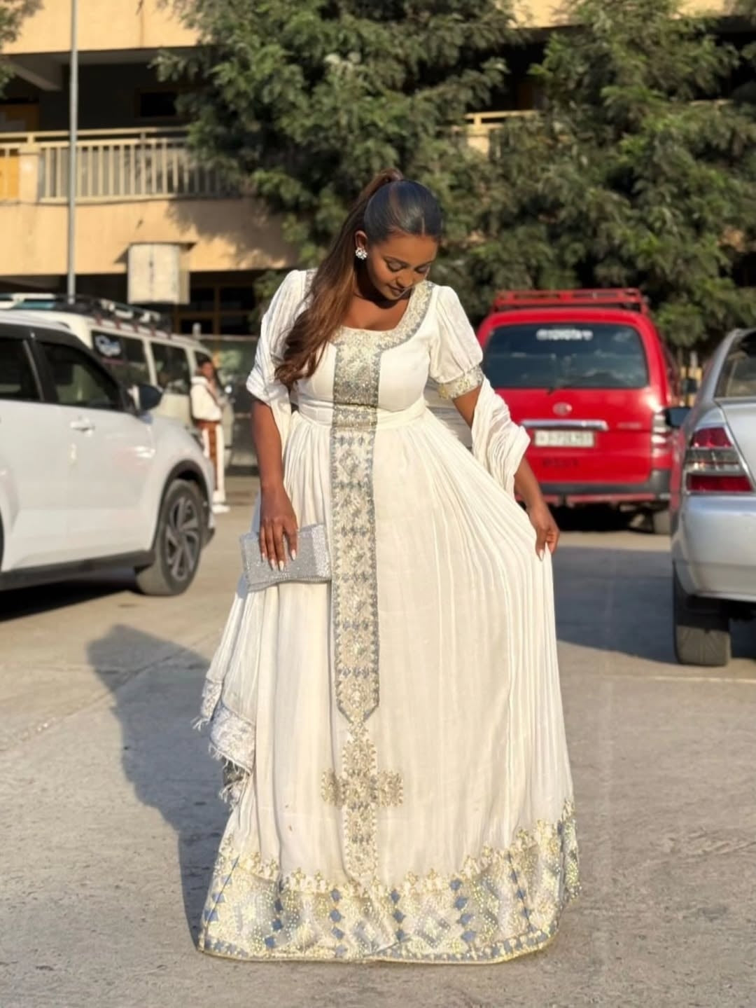 Woman in a white and gold White Ethiopian Traditional Dress  standing in a parking lot with cars and trees in the background.