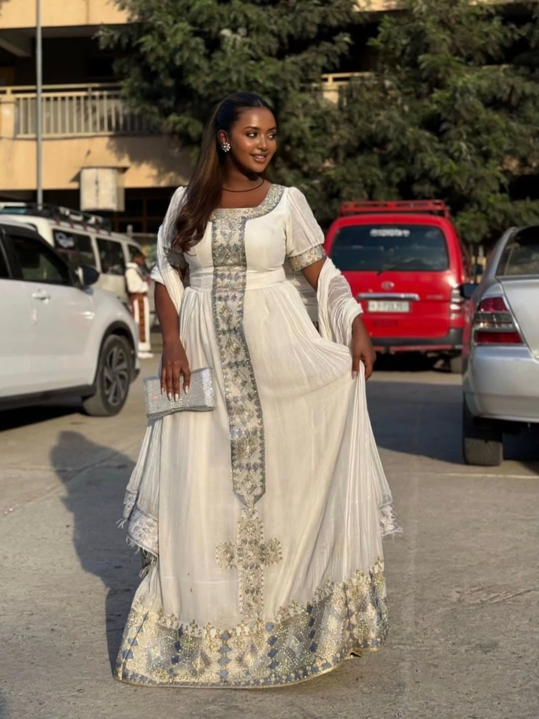 Woman in a white and gray White Ethiopian Traditional Dress  standing in a parking lot with cars and trees in the background.