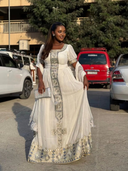 Woman in a white and gray White Ethiopian Traditional Dress  standing in a parking lot with cars and trees in the background.