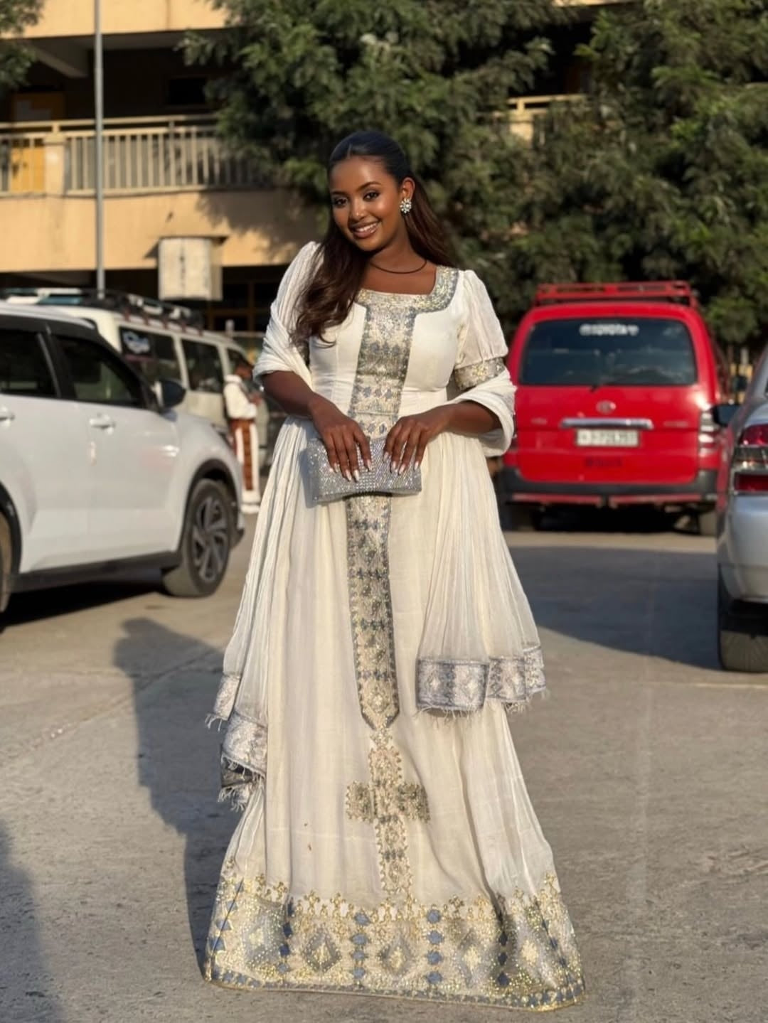 Woman in a white and gold traditional White Ethiopian Traditional Dress  standing on a street with cars and trees in the background.