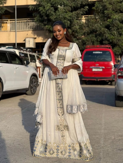 Woman in a white and gold traditional White Ethiopian Traditional Dress  standing on a street with cars and trees in the background.