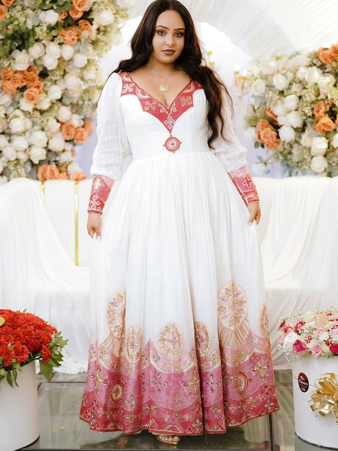Woman in a white and red embroidered red Habesha Kemis standing in front of floral decorations.