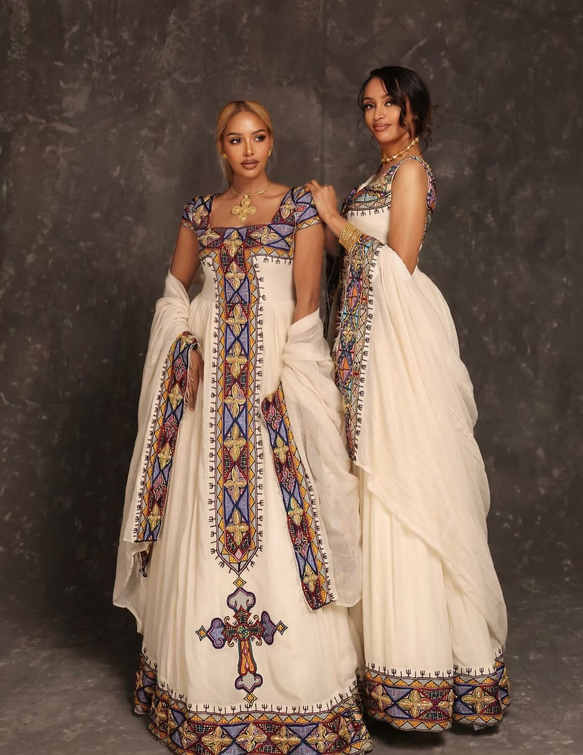 Two women modeling traditional Ethiopian Dress , featuring off-white/cream embroidered dresses with intricate geometric patterns in blue, purple, and gold, set against a dark textured studio backdrop.