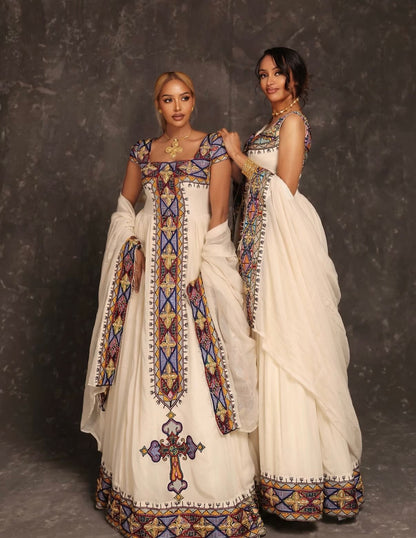 Two women modeling traditional Ethiopian Dress , featuring off-white/cream embroidered dresses with intricate geometric patterns in blue, purple, and gold, set against a dark textured studio backdrop.