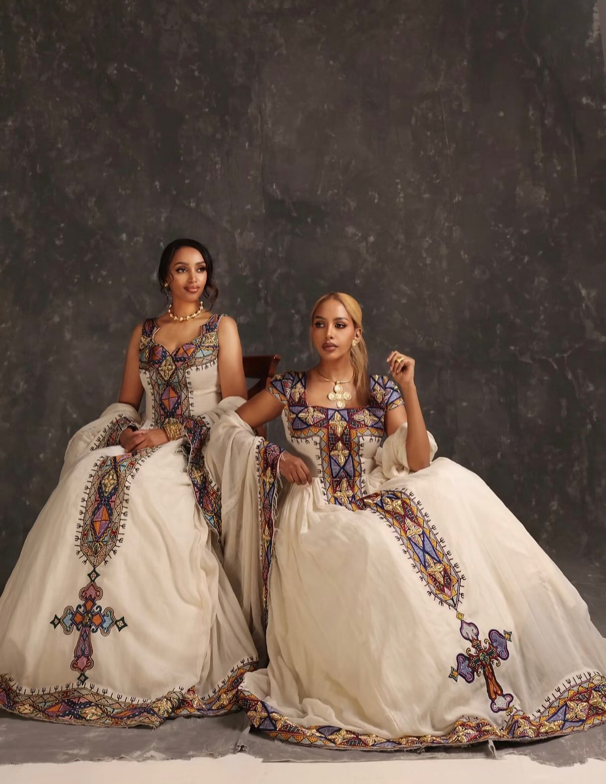 Two women seated side by side in ornate ivory Ethiopian Dress  with colorful embroidered details, posed against a dark textured studio backdrop.