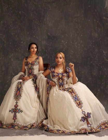 Two women seated side by side in ornate ivory Ethiopian Dress  with colorful embroidered details, posed against a dark textured studio backdrop.