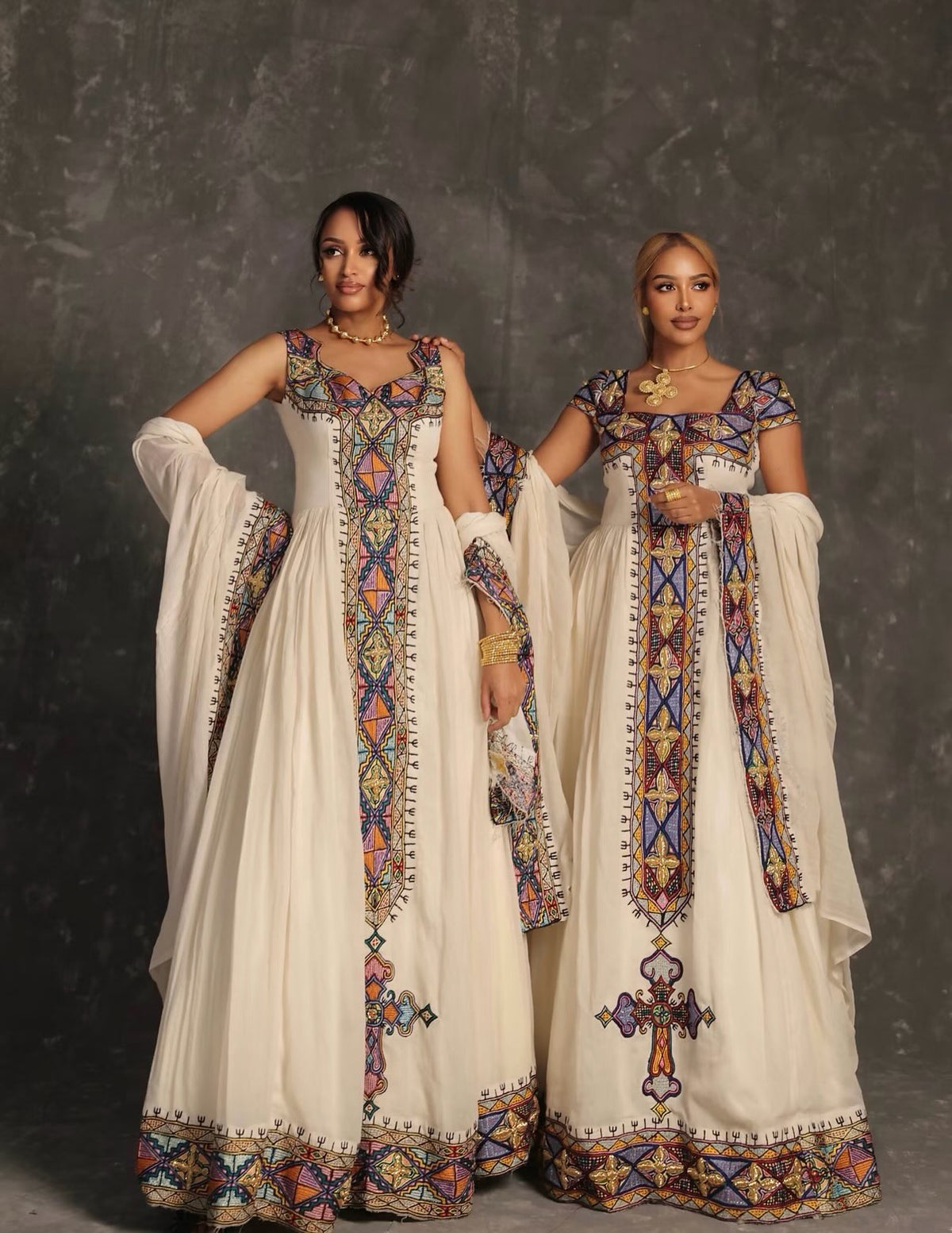 Two women modeling traditional South Asian salwar kameez Ethiopian Dress  with intricate multicolor geometric embroidery on an off-white base, set against a dark textured studio backdrop.