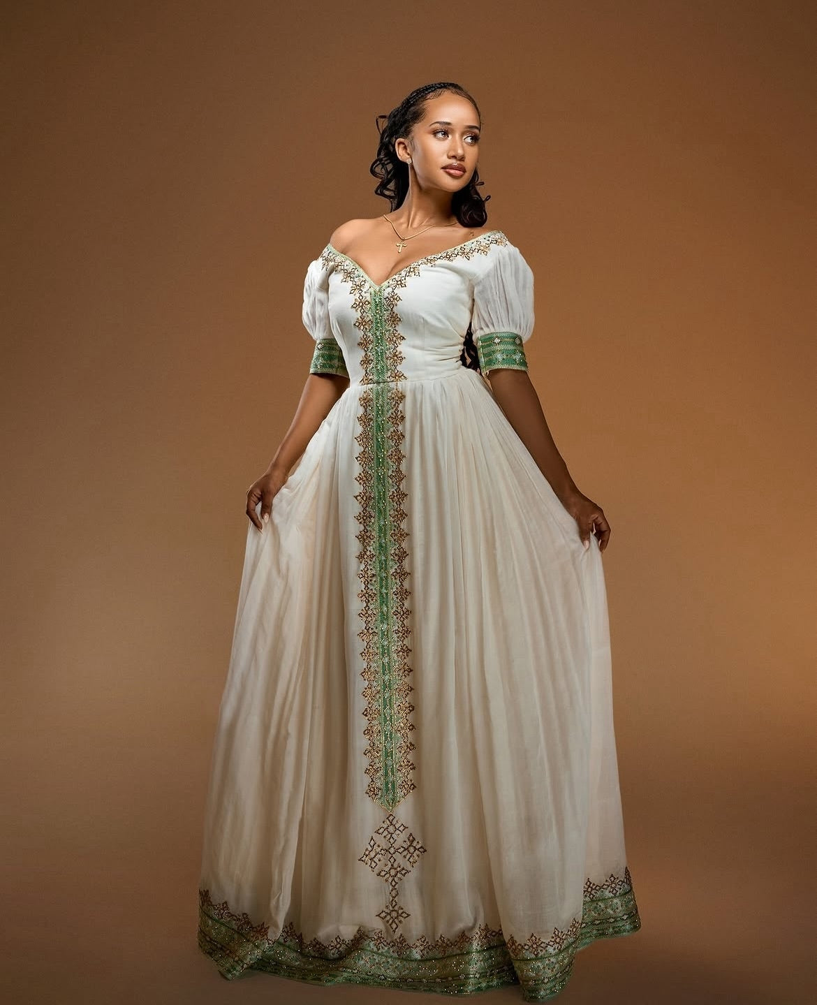 A woman modeling a long off-the-shoulder white Habesha Zuria with green and gold embroidered detailing down the front, on a solid brown studio background.