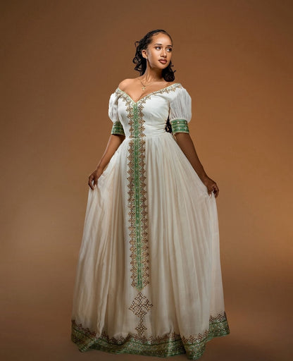 A woman modeling a long off-the-shoulder white Habesha Zuria with green and gold embroidered detailing down the front, on a solid brown studio background.