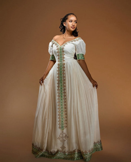 A woman modeling a long off-the-shoulder white Habesha Zuria with green and gold embroidered detailing down the front, on a solid brown studio background.