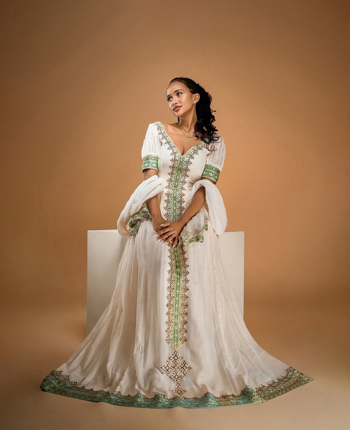 A woman models a white traditional Habesha Zuria with green and gold embroidery, standing on a white pedestal against a warm brown studio background.