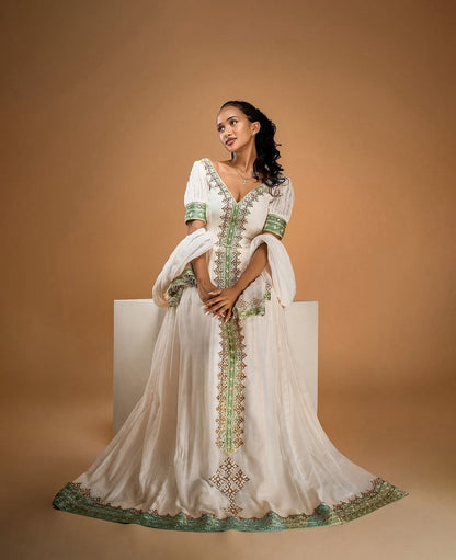 A woman models a white traditional Habesha Zuria with green and gold embroidery, standing on a white pedestal against a warm brown studio background.