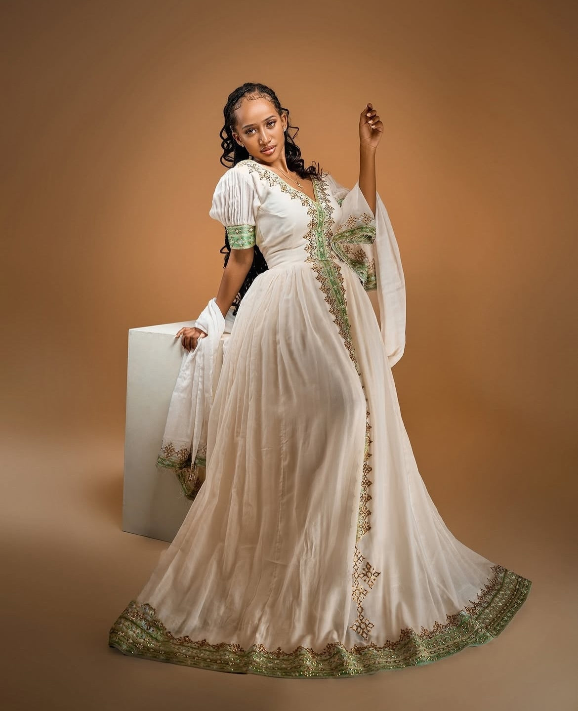 A woman models a traditional long white Habesha Zuria with green embroidered details and a wide green-trimmed hem, posed next to a white pedestal against a warm brown studio backdrop.