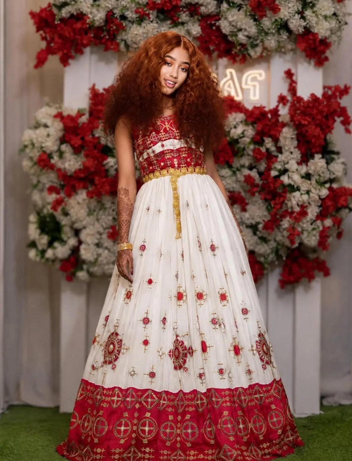 A woman with long, curly red hair models a traditional  Ethiopian dress The dress features a white skirt adorned with red floral motifs and a red border, paired with a red and gold embroidered choli and a gold belt.