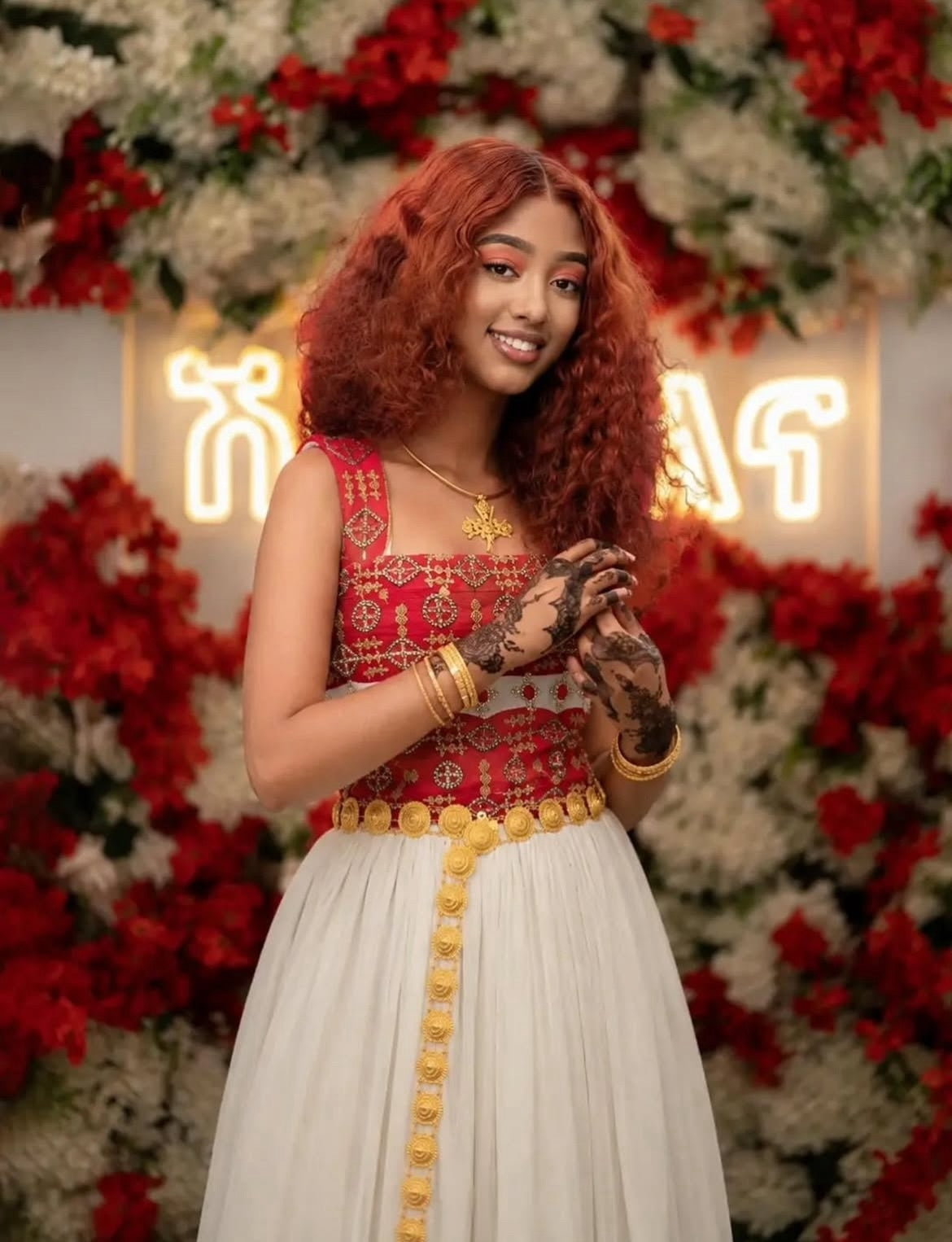 A woman with curly red hair wearing a red and white embroidered traditional-style  Ethiopian dress  with gold jewelry, standing in front of a floral backdrop with red and white flowers and a glowing sign.