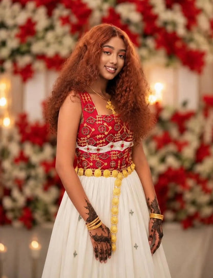 A woman with long, curly auburn hair is wearing a traditional  Ethiopian dress  featuring a red embroidered bodice, a white skirt, and a yellow beaded waist belt. She is standing in front of a decorative floral backdrop with red and white flowers and warm string lights.