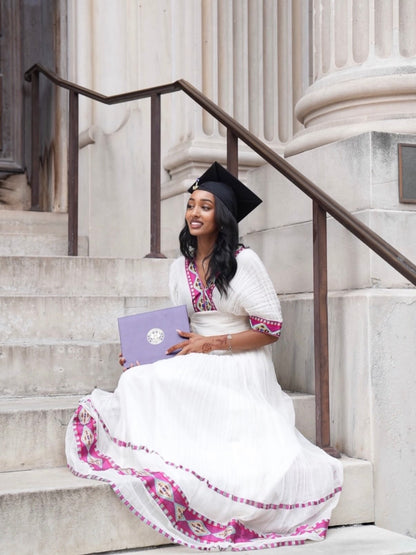 A woman in a white graduation Habesha kemis with pink and purple embroidery sits on stone steps holding a purple diploma folder, wearing a black cap.