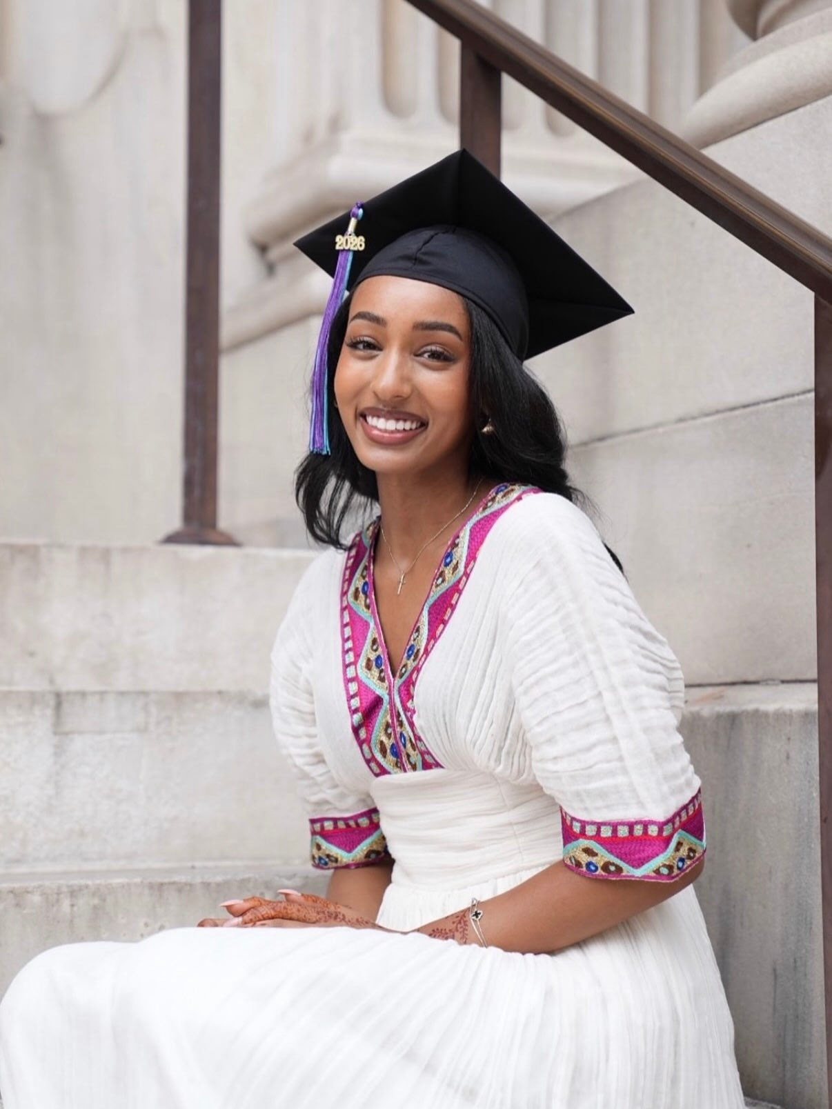 A smiling woman wearing a black graduation Habesha kemis sits on stone steps, dressed in a white traditional outfit with bright pink and multicolor embroidered trim.