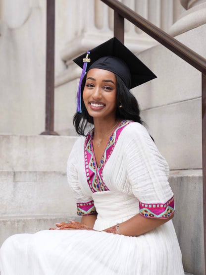 A smiling woman wearing a black graduation Habesha kemis sits on stone steps, dressed in a white traditional outfit with bright pink and multicolor embroidered trim.