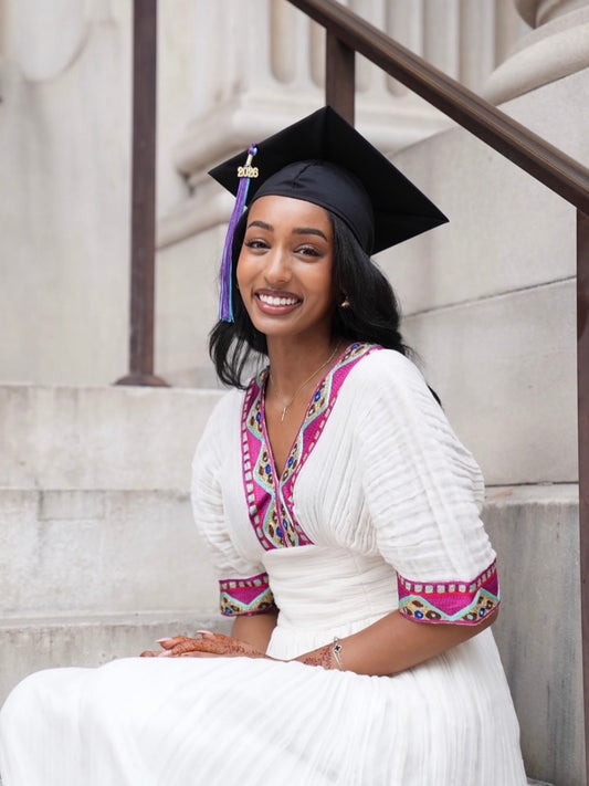 A smiling woman wearing a black graduation Habesha kemis sits on stone steps, dressed in a white traditional outfit with bright pink and multicolor embroidered trim.