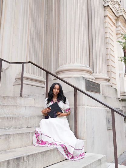 A woman sits on the stone steps of a grand neoclassical building with large columns, wearing a white traditional-style Habesha kemis with pink and purple embroidered borders and holding a black book.