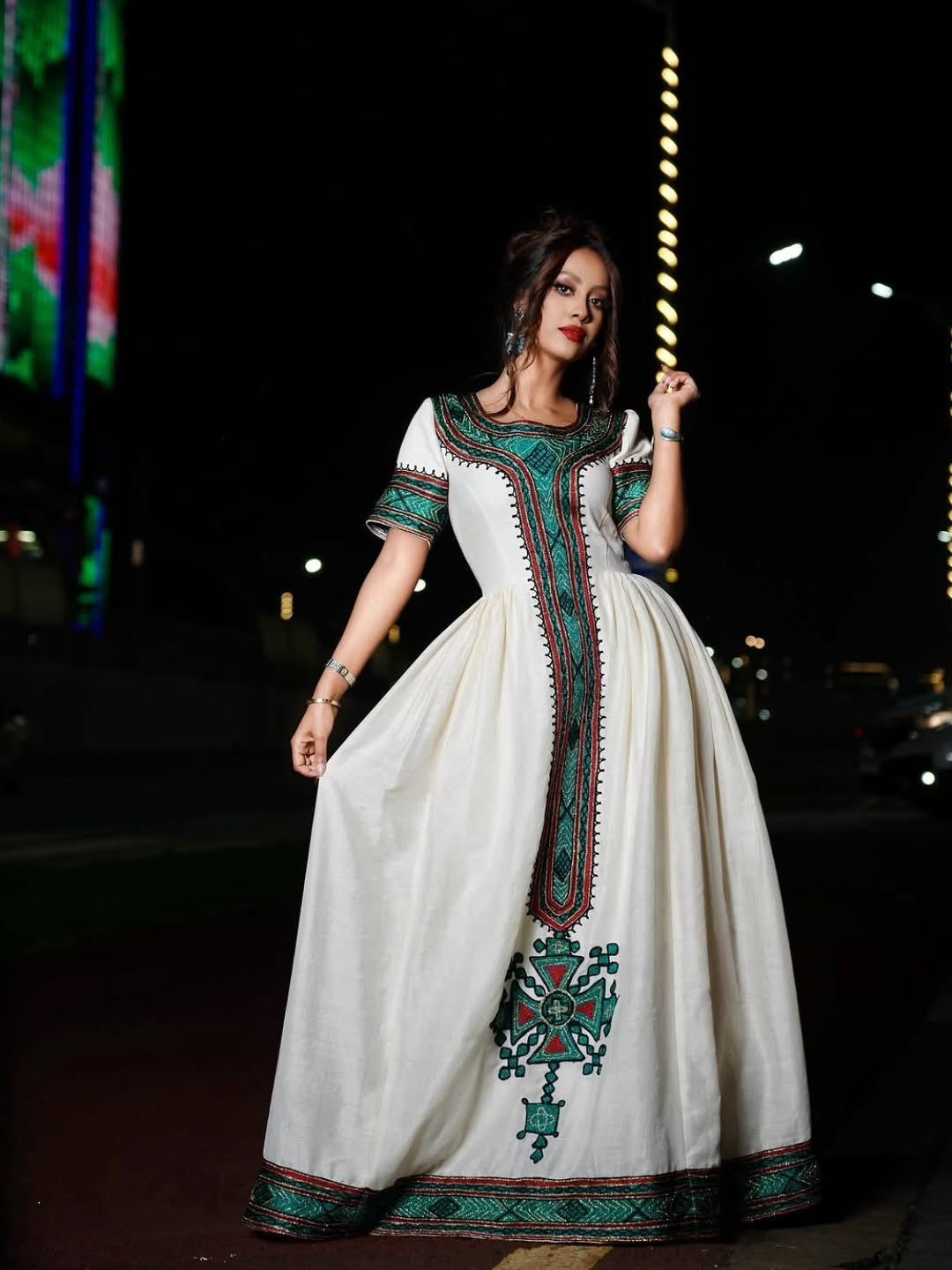A woman wearing a long white dress with green and red embroidered details, standing outdoors at night with blurred city lights in the background.
