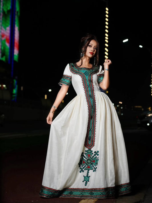 A woman wearing a long white dress with green and red embroidered details, standing outdoors at night with blurred city lights in the background.
