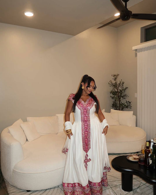 A woman wearing a white traditional Habesha Kemis with pink and red embroidery stands in a living room in front of a curved white sofa.