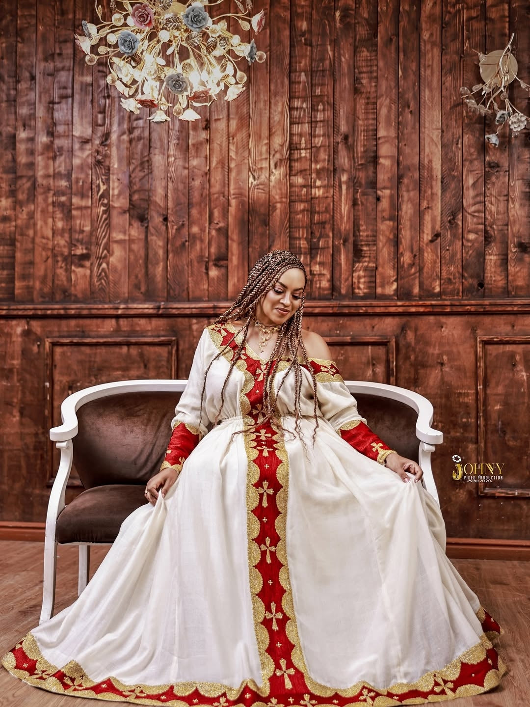 Woman in a white and red Ethiopian Dress with gold accents sitting on a chair against a wooden paneled wall.