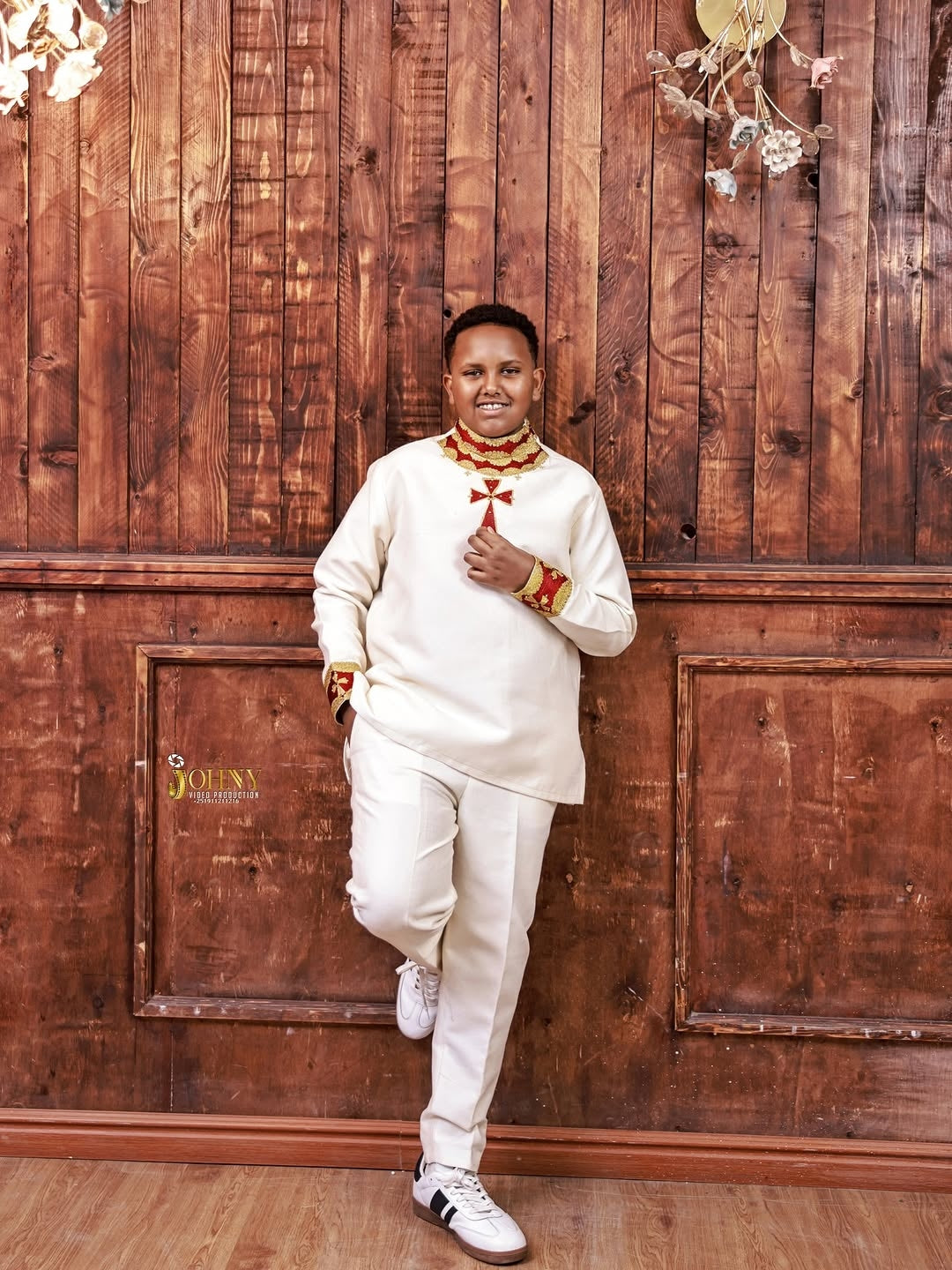 Person in a white outfit with red accents standing against a wooden paneled wall.